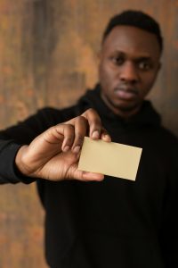 Soft focus of African American male in black casual clothes looking at camera while showing yellow blank business card in hand